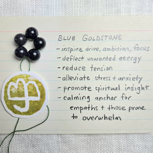 A handmade blue glass bead mala necklace with a gold-toned circular pendant featuring a white symbol, displayed on a handwritten note that lists the benefits of a blue gemstone.