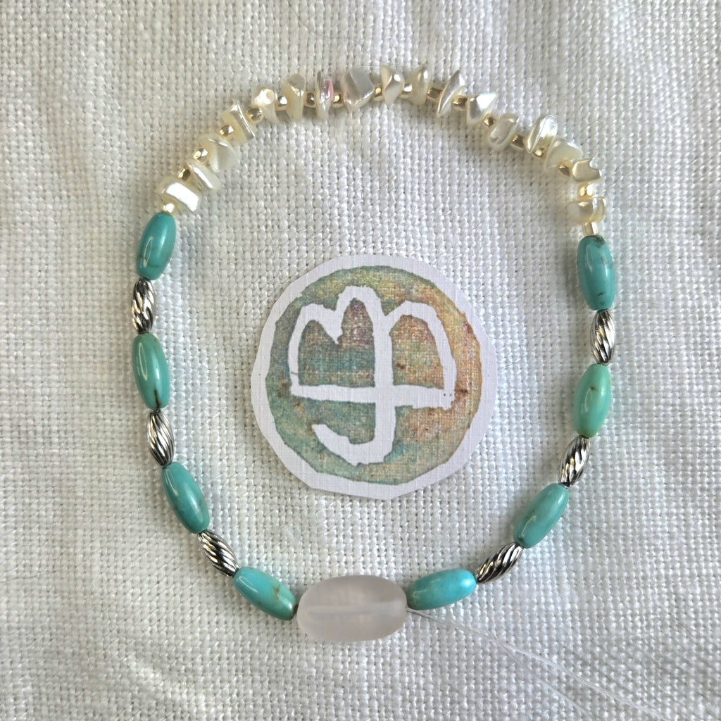 A handmade beaded bracelet with turquoise-blue oval beads, silver-toned twisted rope beads, and a large milky white oval bead, displayed on a textured white fabric next to a circular patch with a white symbol.