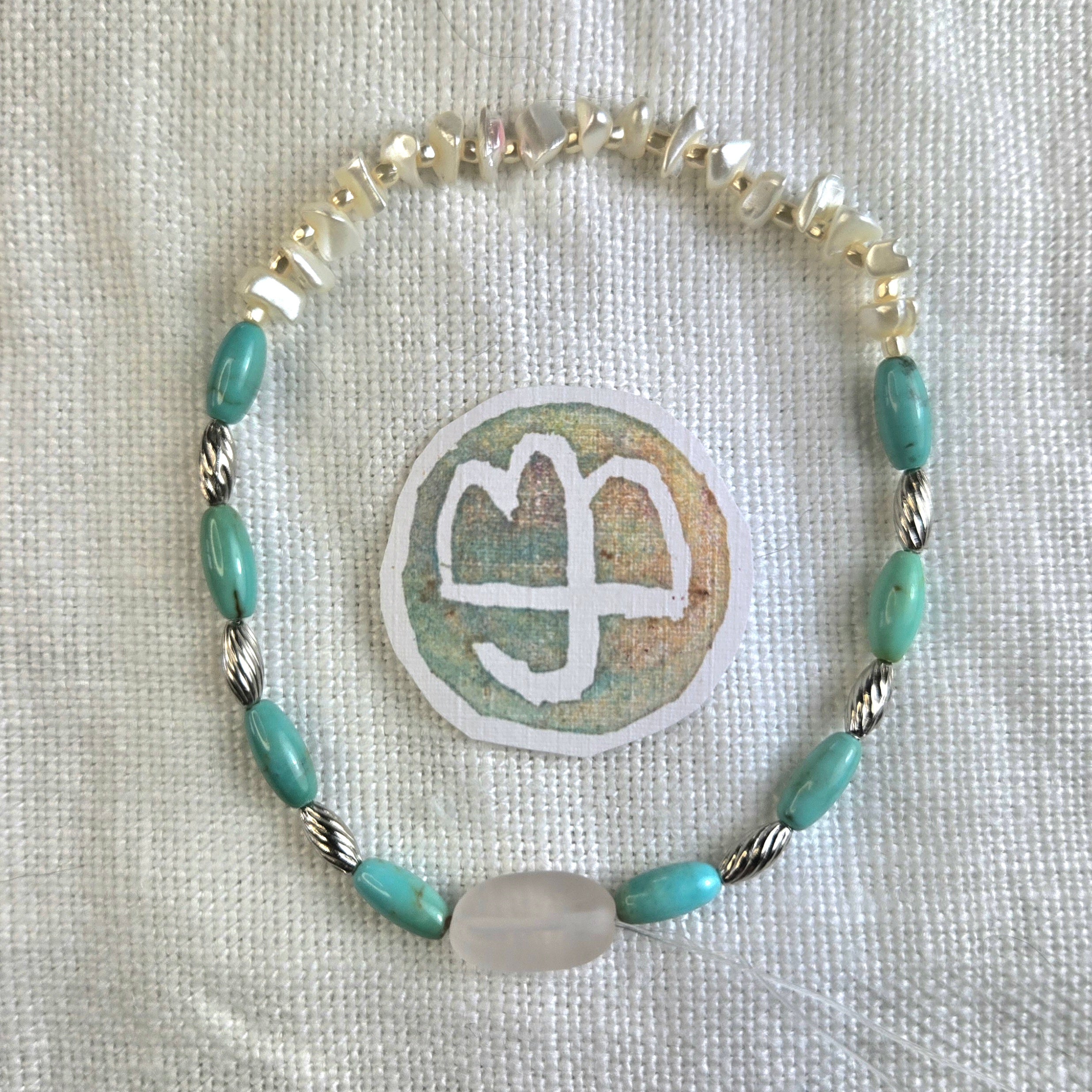 A handmade beaded bracelet with turquoise-blue oval beads, silver-toned twisted rope beads, and a large milky white oval bead, displayed on a textured white fabric next to a circular patch with a white symbol.