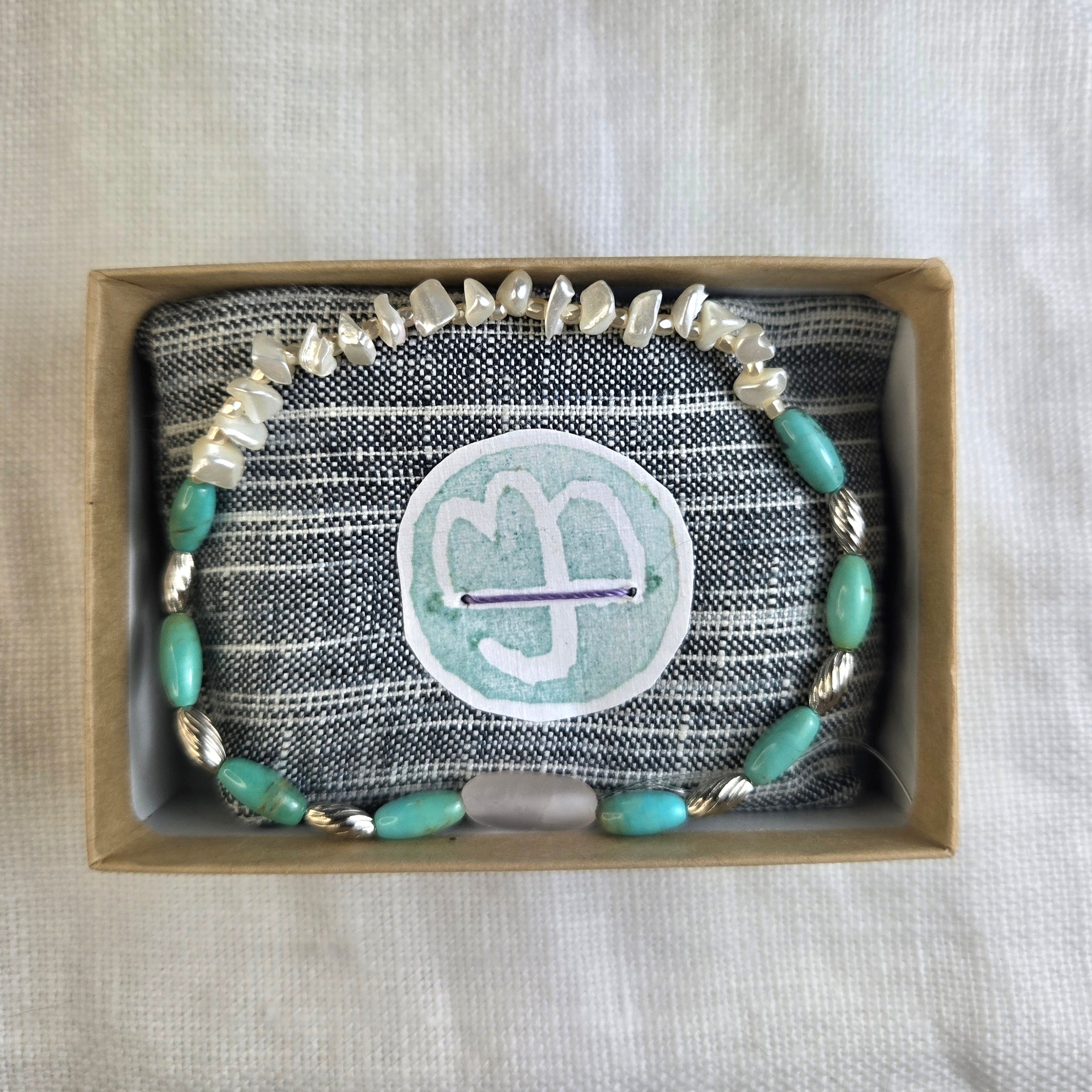 A handmade beaded bracelet with turquoise-green beads, silver-toned chunky stones, and twisted silver spacers, displayed inside a brown gift box on a gray striped fabric cushion with a custom round sticker.
