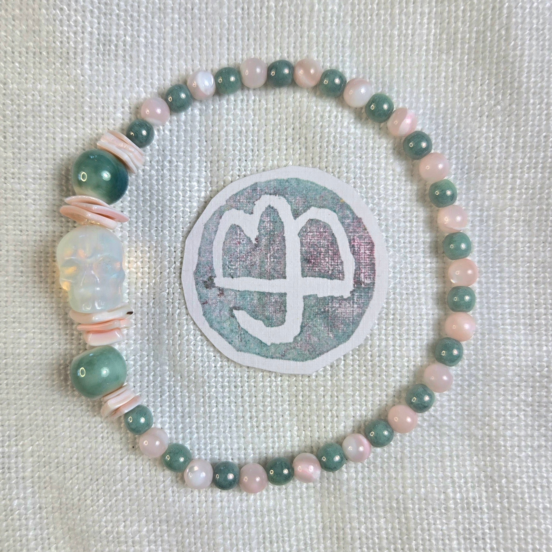 A beaded bracelet arranged in a circle on a textured off-white fabric background, featuring light green and pale pink beads, a translucent white carved bead, and thin pink spacer beads. A circular sticker with a white symbol is placed behind the bracelet.