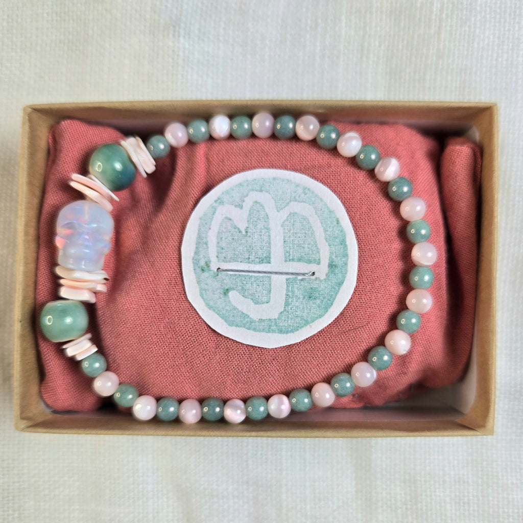 A beaded bracelet with pastel pink, sage green, and white beads, displayed inside a brown gift box on a dusty pink fabric insert. The center of the insert features a circular white patch with a white monogram logo.