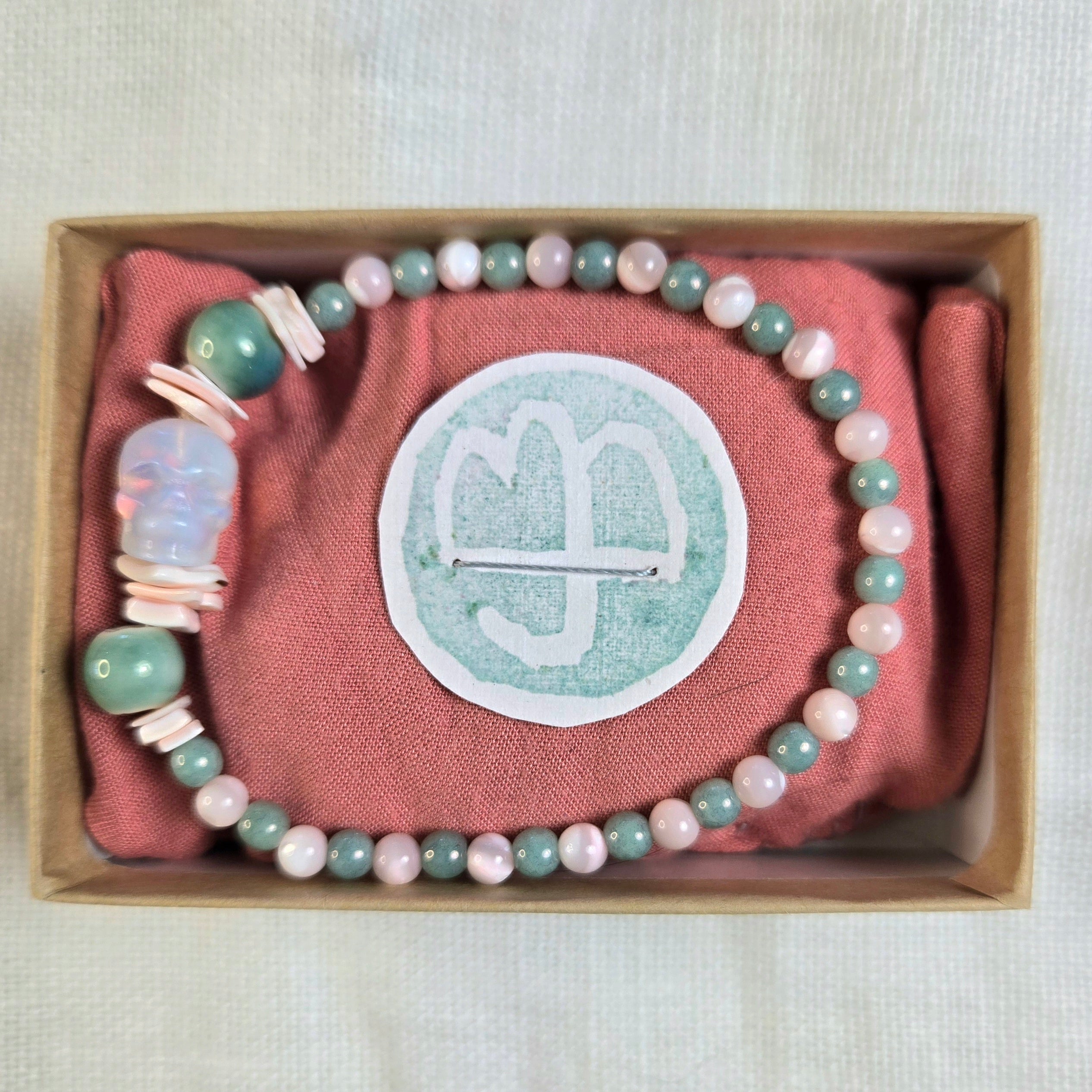 A beaded bracelet with pastel pink, sage green, and white beads, displayed inside a brown gift box on a dusty pink fabric insert. The center of the insert features a circular white patch with a white monogram logo.