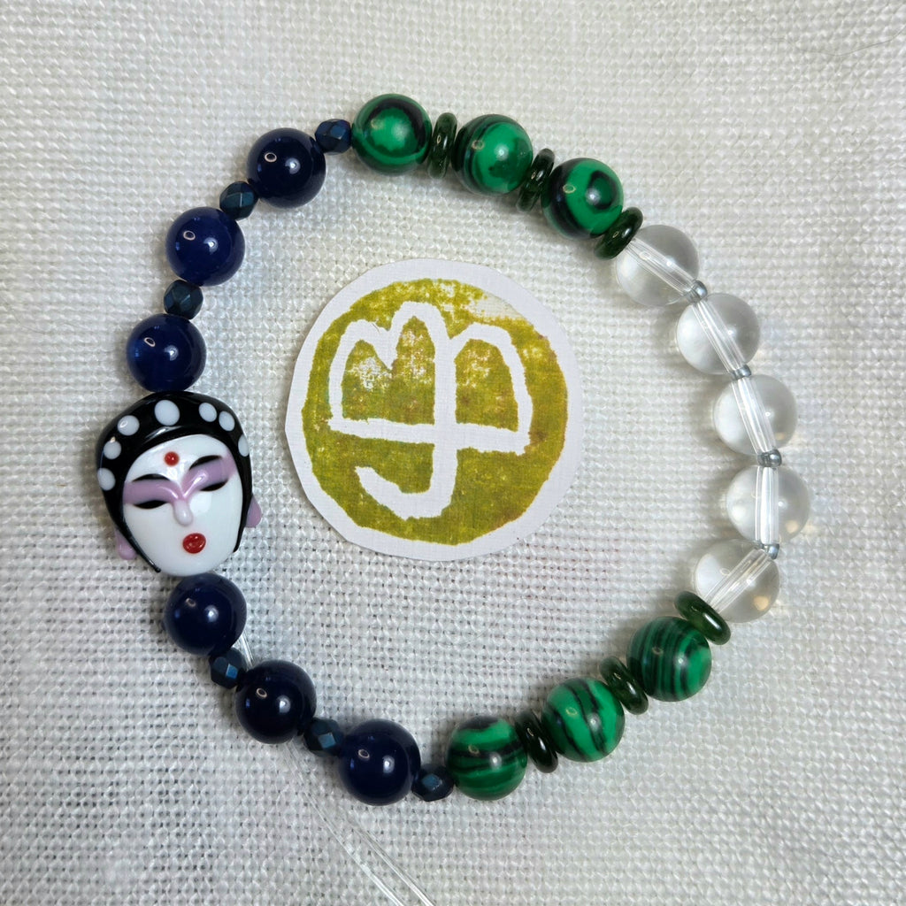 A beaded bracelet arranged in a circle on a white textured fabric background. The bracelet features dark navy blue beads, green marbled beads, clear round beads, and a decorative black-and-white porcelain-style face charm with red and purple details.