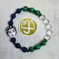 A beaded bracelet arranged in a circle on a white textured fabric background. The bracelet features dark navy blue beads, green marbled beads, clear round beads, and a decorative black-and-white porcelain-style face charm with red and purple details.