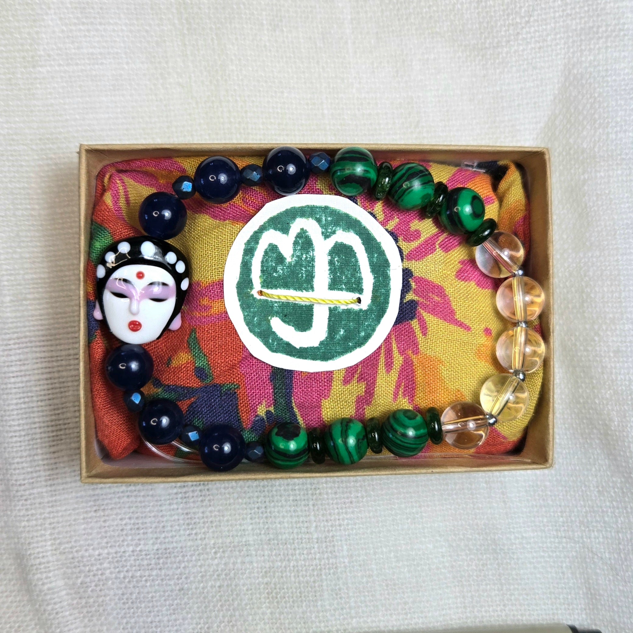 A beaded bracelet or necklace set displayed in a small kraft box on a patterned fabric insert. The set includes glossy dark blue beads, green marbled beads, clear faceted beads, a white and black painted face charm, and a round white sticker with a green monogram.