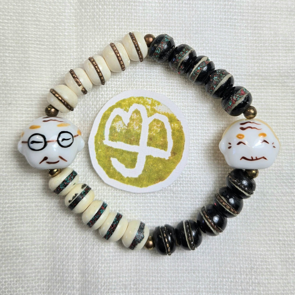 A handmade beaded stretch bracelet featuring black and cream beads, striped accents, and two white ceramic head charms with painted facial features. A round gold and white logo is placed in the center of the bracelet on a textured white fabric background.