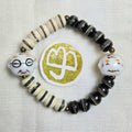 A handmade beaded stretch bracelet featuring black and cream beads, striped accents, and two white ceramic head charms with painted facial features. A round gold and white logo is placed in the center of the bracelet on a textured white fabric background.
