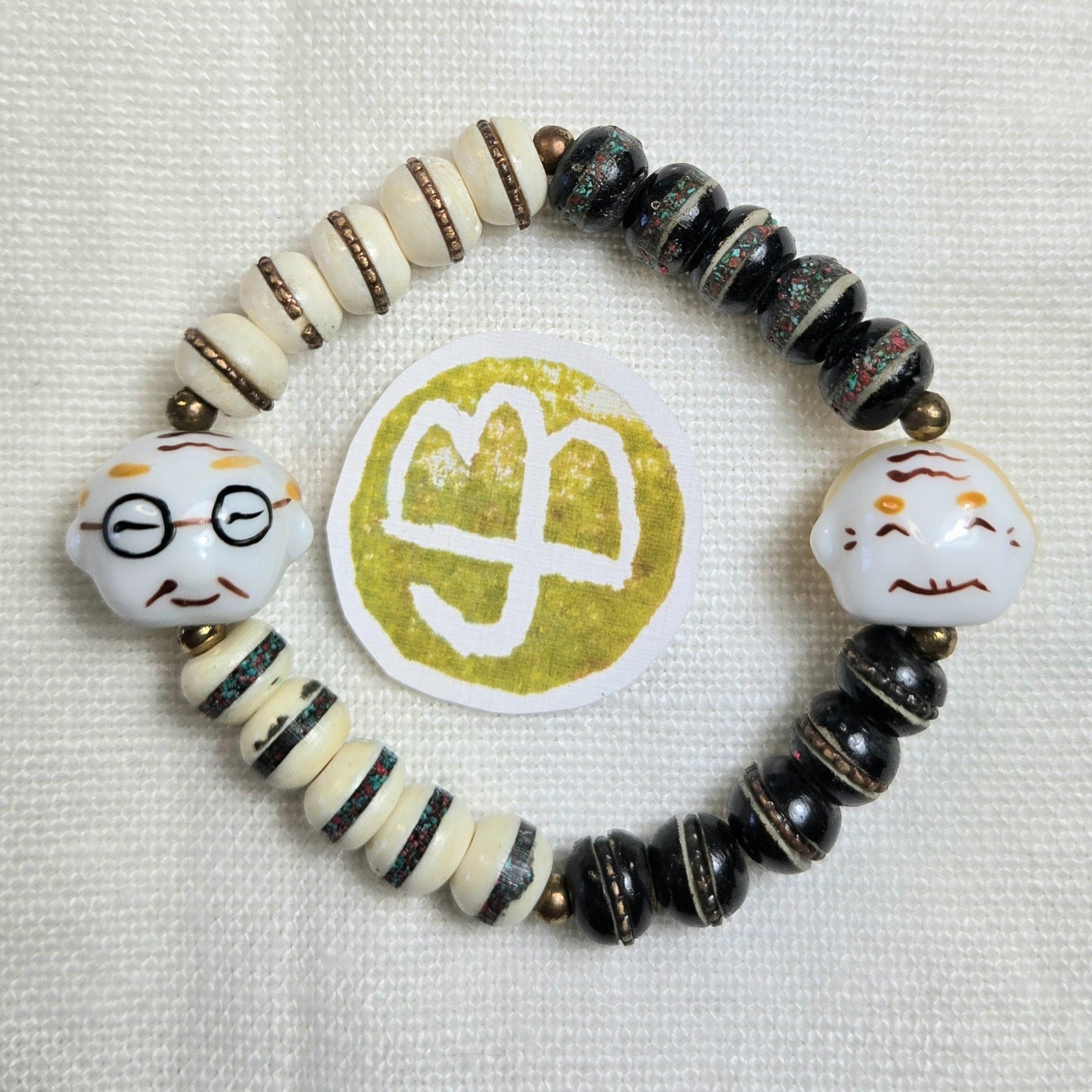 A handmade beaded stretch bracelet featuring black and cream beads, striped accents, and two white ceramic head charms with painted facial features. A round gold and white logo is placed in the center of the bracelet on a textured white fabric background.