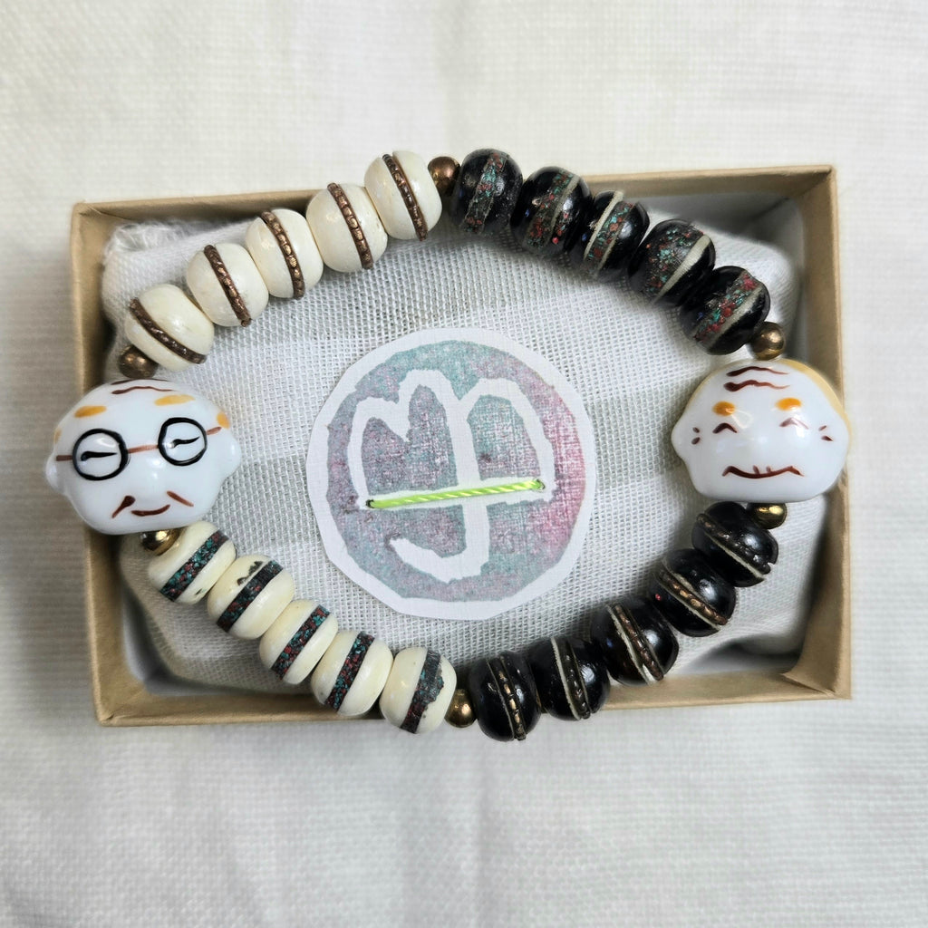 A beaded bracelet displayed inside a small kraft gift box on a white fabric surface. The bracelet features glossy black and cream beads, striped cream-and-black beads, and two white character charms with painted facial features.