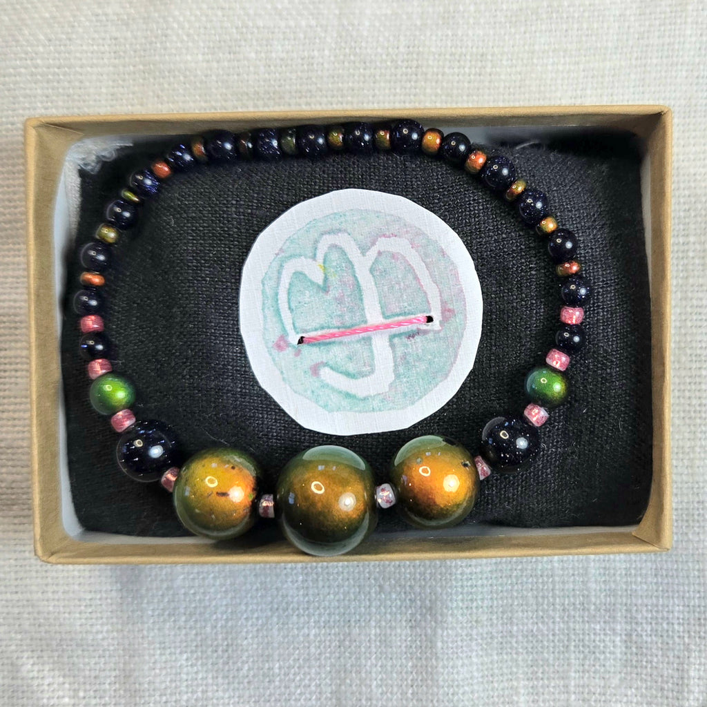 A beaded bracelet displayed inside a light wood jewelry box on a black fabric insert. The bracelet features large polished gold-toned beads at the front, dark black beads, and smaller multicolored beads including green, pink, and brown.