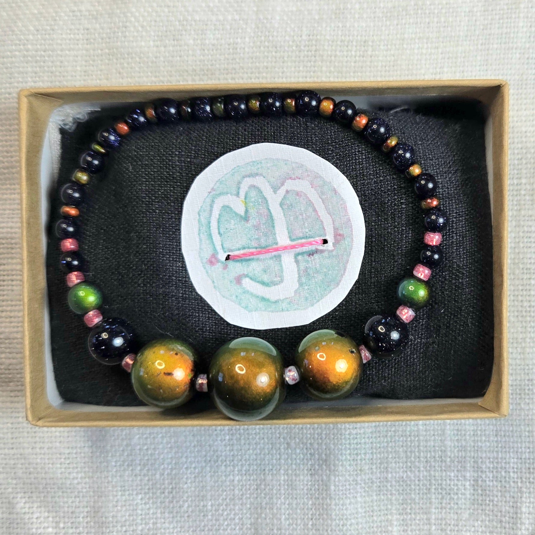A beaded bracelet displayed inside a light wood jewelry box on a black fabric insert. The bracelet features large polished gold-toned beads at the front, dark black beads, and smaller multicolored beads including green, pink, and brown.
