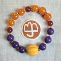 A beaded stretch bracelet arranged in a circle on an off-white linen fabric background, featuring orange, purple, yellow, and reddish-brown beads separated by small gold spacer beads. A round brown patch with a logo is placed behind the bracelet.
