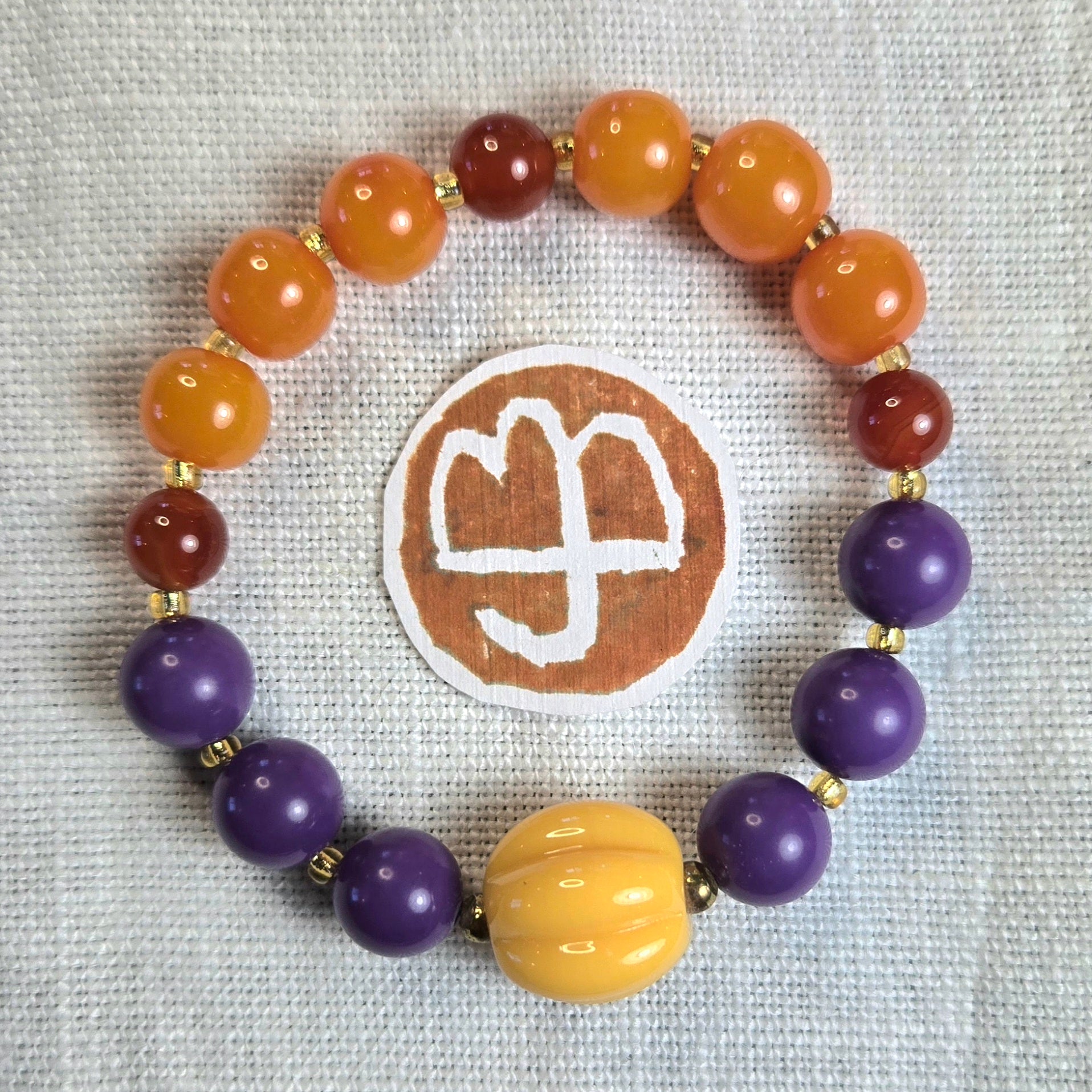 A beaded stretch bracelet arranged in a circle on an off-white linen fabric background, featuring orange, purple, yellow, and reddish-brown beads separated by small gold spacer beads. A round brown patch with a logo is placed behind the bracelet.