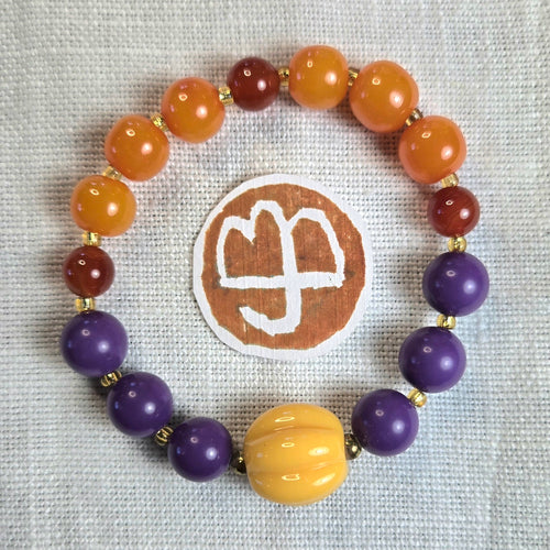 A beaded stretch bracelet arranged in a circle on an off-white linen fabric background, featuring orange, purple, yellow, and reddish-brown beads separated by small gold spacer beads. A round brown patch with a logo is placed behind the bracelet.