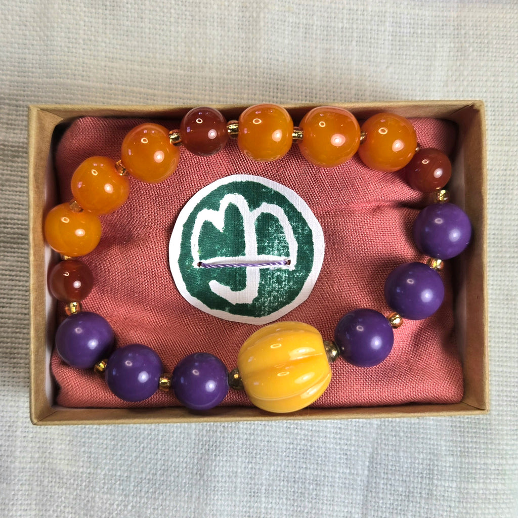 A beaded bracelet displayed inside a small kraft gift box lined with pink fabric. The bracelet features glossy orange and brown beads, several purple beads, a large yellow bead at the front, and small gold spacer beads.