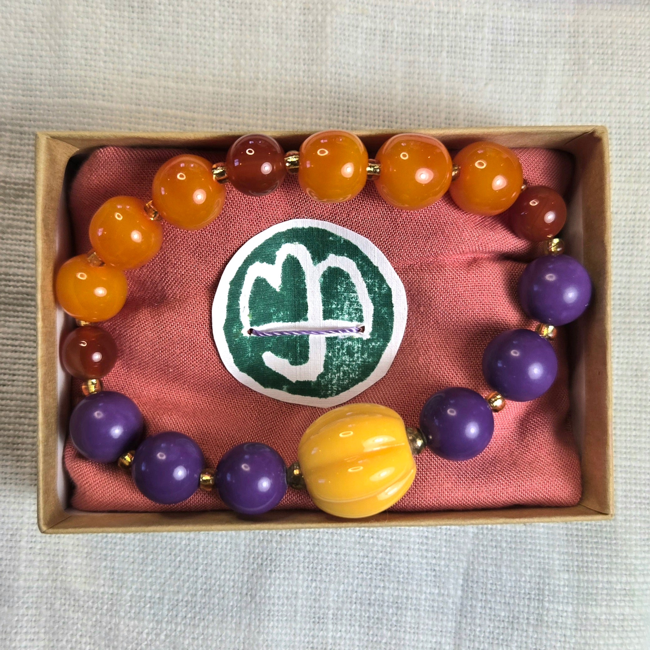 A beaded bracelet displayed inside a small kraft gift box lined with pink fabric. The bracelet features glossy orange and brown beads, several purple beads, a large yellow bead at the front, and small gold spacer beads.
