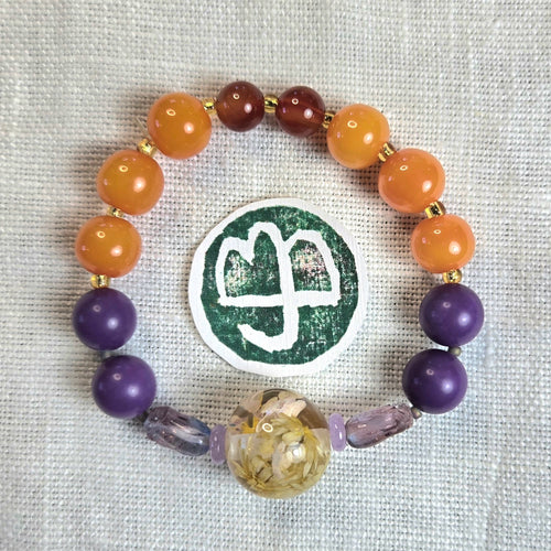 A beaded stretch bracelet arranged in a circle on a textured white fabric background, featuring orange and purple beads, gold spacer beads, and a large decorative central bead with dried botanicals. A round green and white sticker is placed in front of the bracelet.