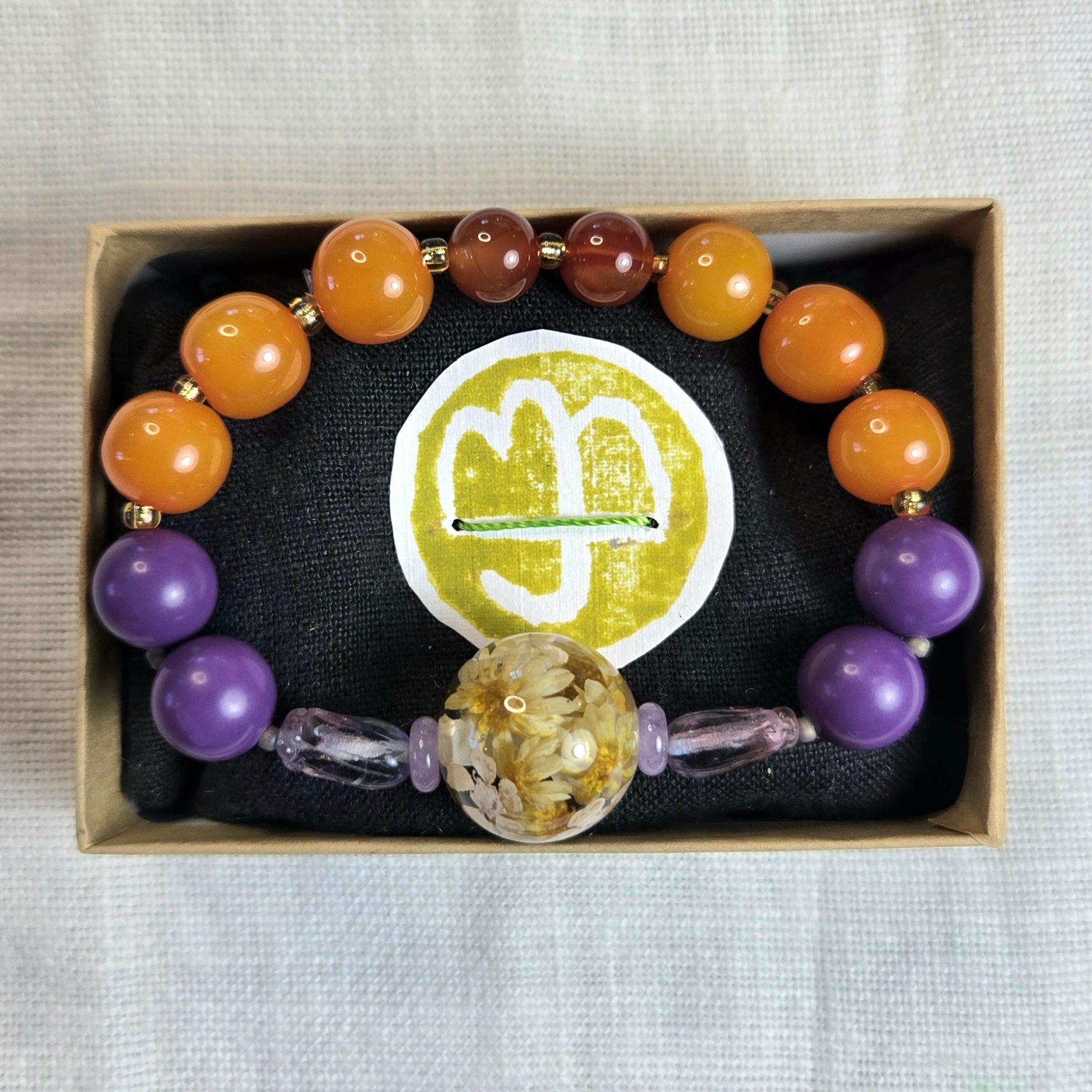 A beaded bracelet displayed inside a small kraft gift box on a black cushion. The bracelet features round beads in orange, brown, and purple, separated by small gold spacer beads, with two clear faceted beads and a large central clear bead filled with tiny flowers.