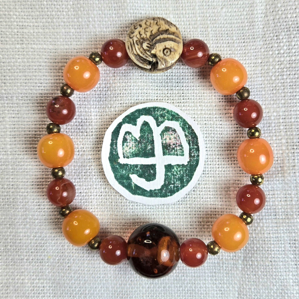 A beaded bracelet arranged in a circle on a textured off-white fabric background. The bracelet features round beads in orange, reddish-brown, and translucent dark brown, separated by small metallic bronze beads.