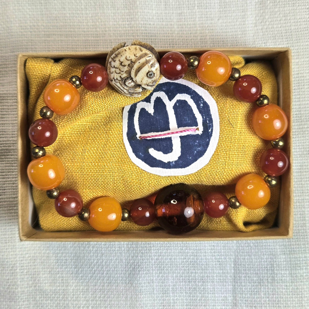A beaded bracelet with orange, reddish-brown, and dark brown polished beads, small metallic spacer beads, and a carved animal-head charm, displayed inside a light wood box lined with yellow fabric.