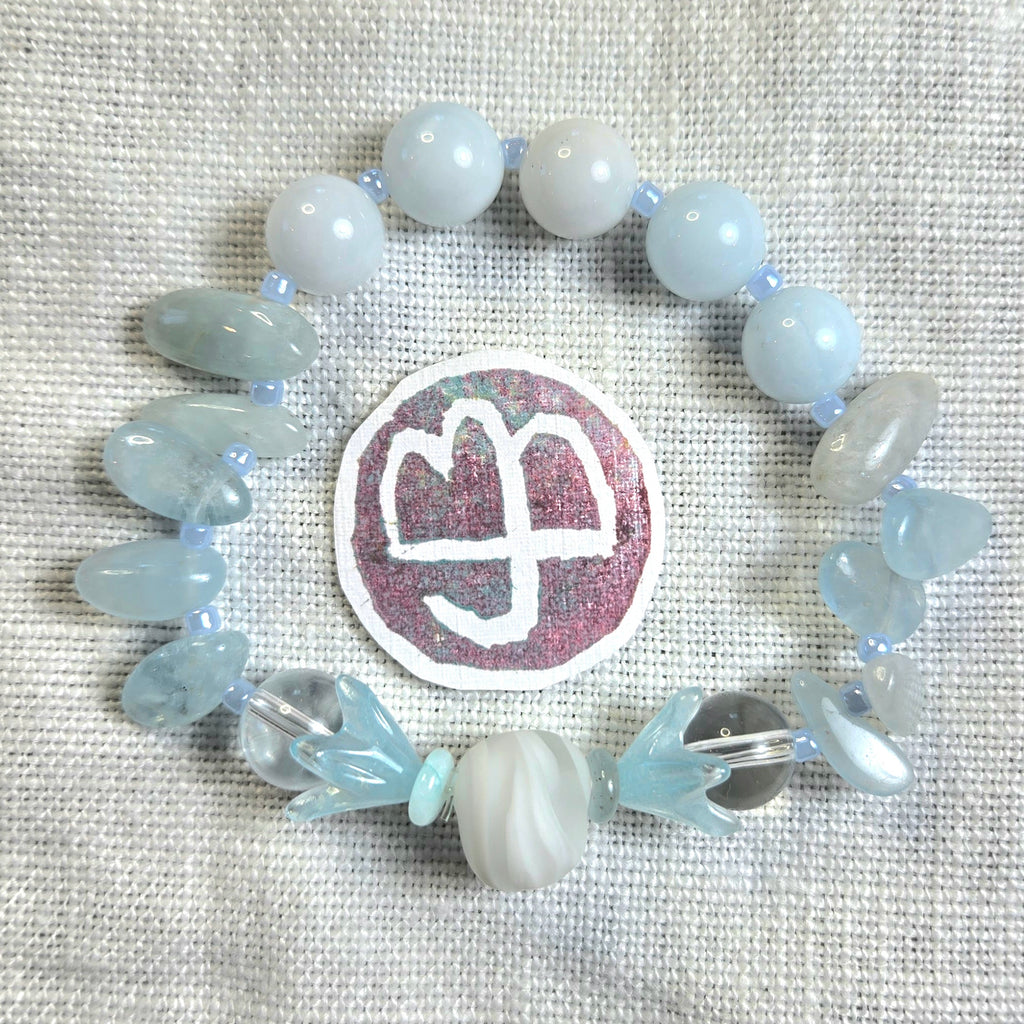 A beaded stretch bracelet arranged in a circle on a textured off-white fabric background. The bracelet features pale blue and white beads, including smooth round beads, polished stone chips, translucent clear beads, and a large central white-and-pink circular charm with a logo.