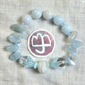 A beaded stretch bracelet arranged in a circle on a textured off-white fabric background. The bracelet features pale blue and white beads, including smooth round beads, polished stone chips, translucent clear beads, and a large central white-and-pink circular charm with a logo.