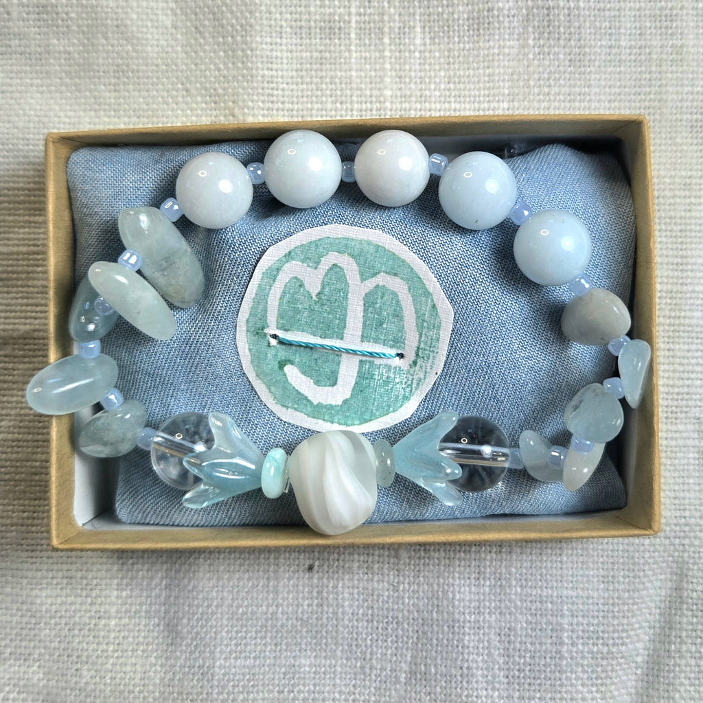 A beaded bracelet with white, pale blue, and gray translucent beads, displayed inside a light wood box lined with gray fabric and a circular mint-green logo patch.