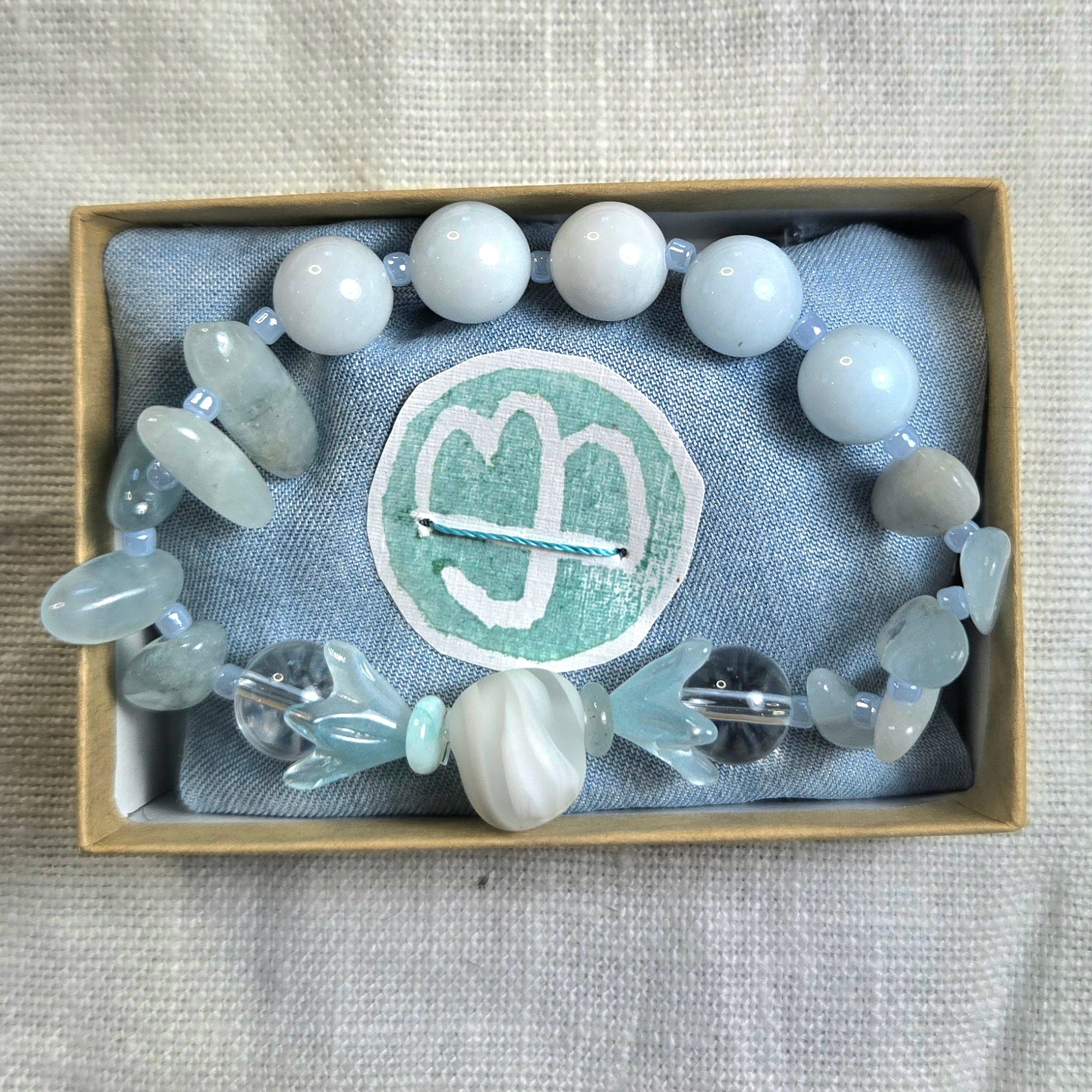 A beaded bracelet with white, pale blue, and gray translucent beads, displayed inside a light wood box lined with gray fabric and a circular mint-green logo patch.