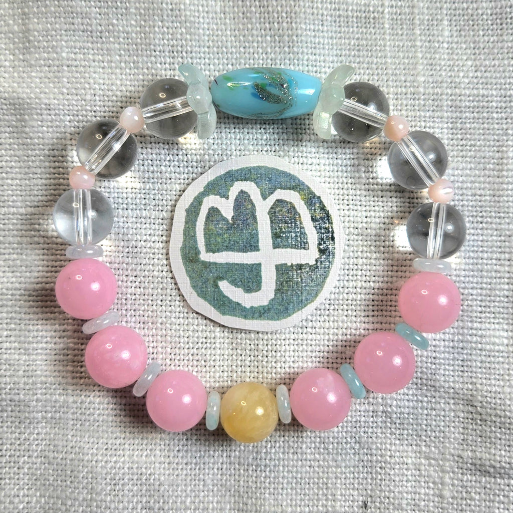 A beaded bracelet arranged in a circle on a textured light beige fabric background, featuring pink round beads, clear faceted beads, a light blue teardrop bead, a pale yellow round bead, and a central round patch with a white symbol.