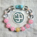 A beaded bracelet arranged in a circle on a textured light beige fabric background, featuring pink round beads, clear faceted beads, a light blue teardrop bead, a pale yellow round bead, and a central round patch with a white symbol.