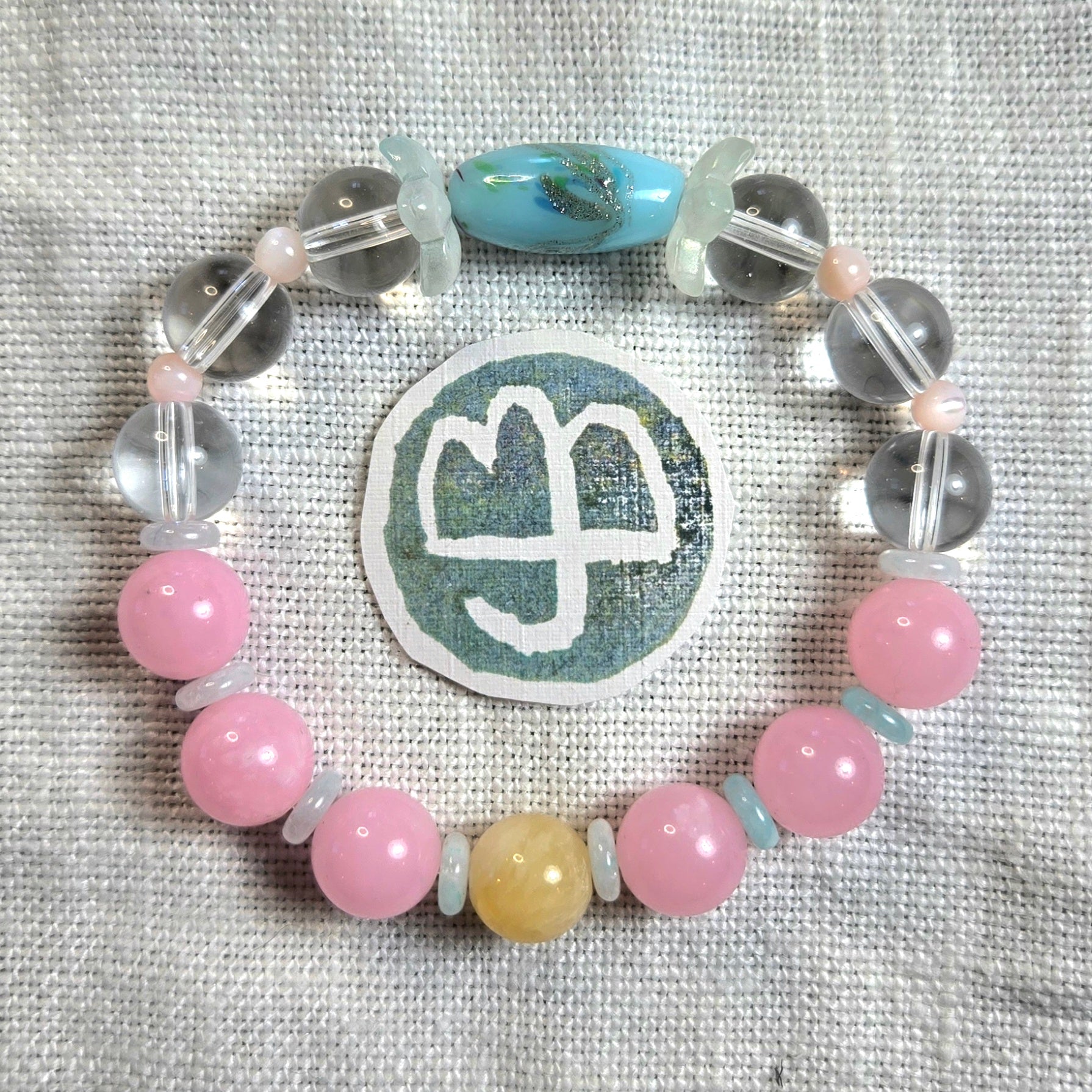 A beaded bracelet arranged in a circle on a textured light beige fabric background, featuring pink round beads, clear faceted beads, a light blue teardrop bead, a pale yellow round bead, and a central round patch with a white symbol.