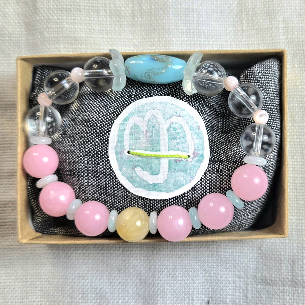 A beaded bracelet displayed inside a small kraft gift box on a dark textured fabric lining. The bracelet features light pink round beads, pale blue translucent beads, clear faceted beads, and one larger light blue oval bead.