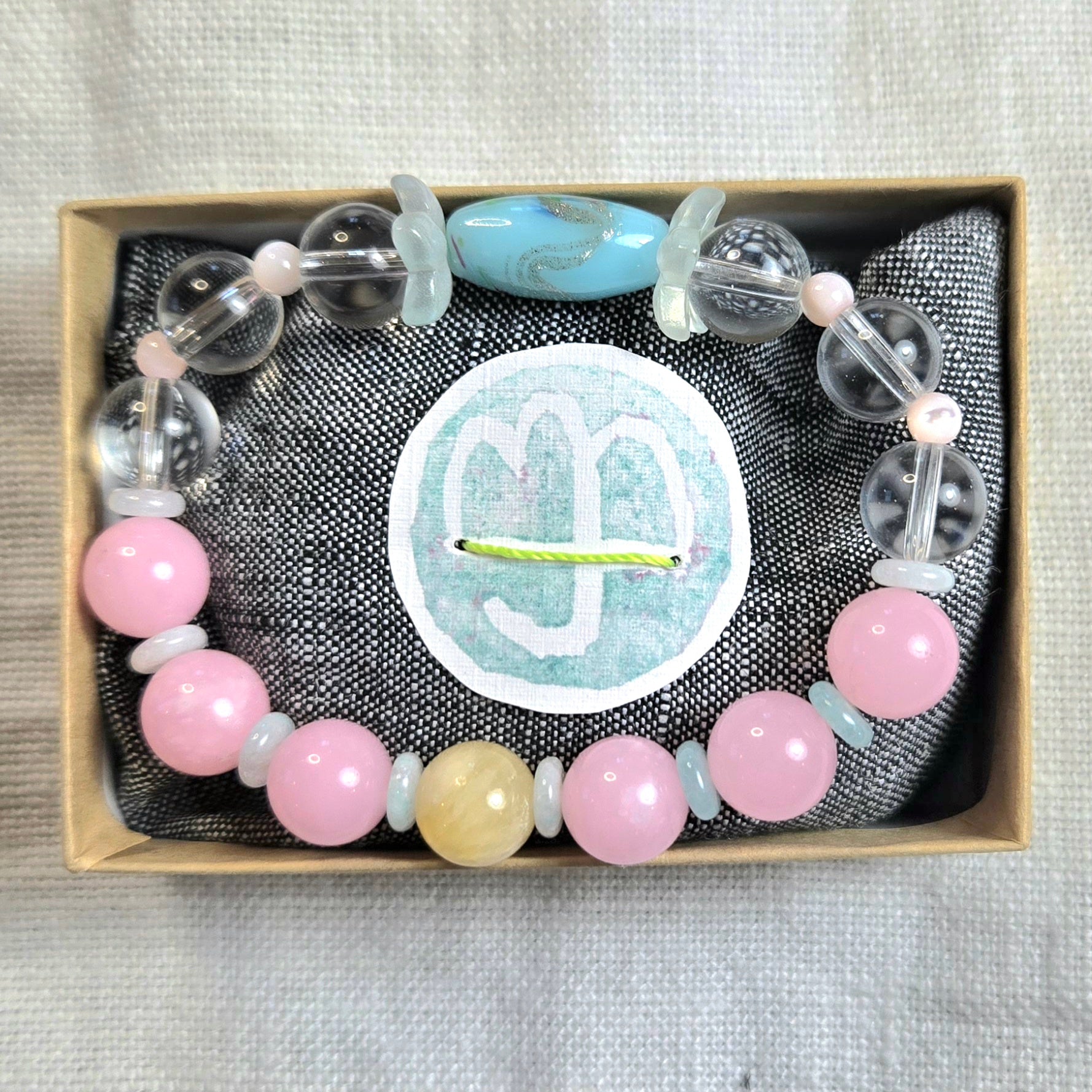 A beaded bracelet displayed inside a small kraft gift box on a dark textured fabric lining. The bracelet features light pink round beads, pale blue translucent beads, clear faceted beads, and one larger light blue oval bead.