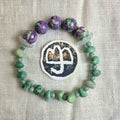 A handmade beaded bracelet arranged in a circle on a textured off-white fabric background. The bracelet features purple and green marbled beads at the top, light green smooth beads, and a central white circular charm with a dark blue symbol.