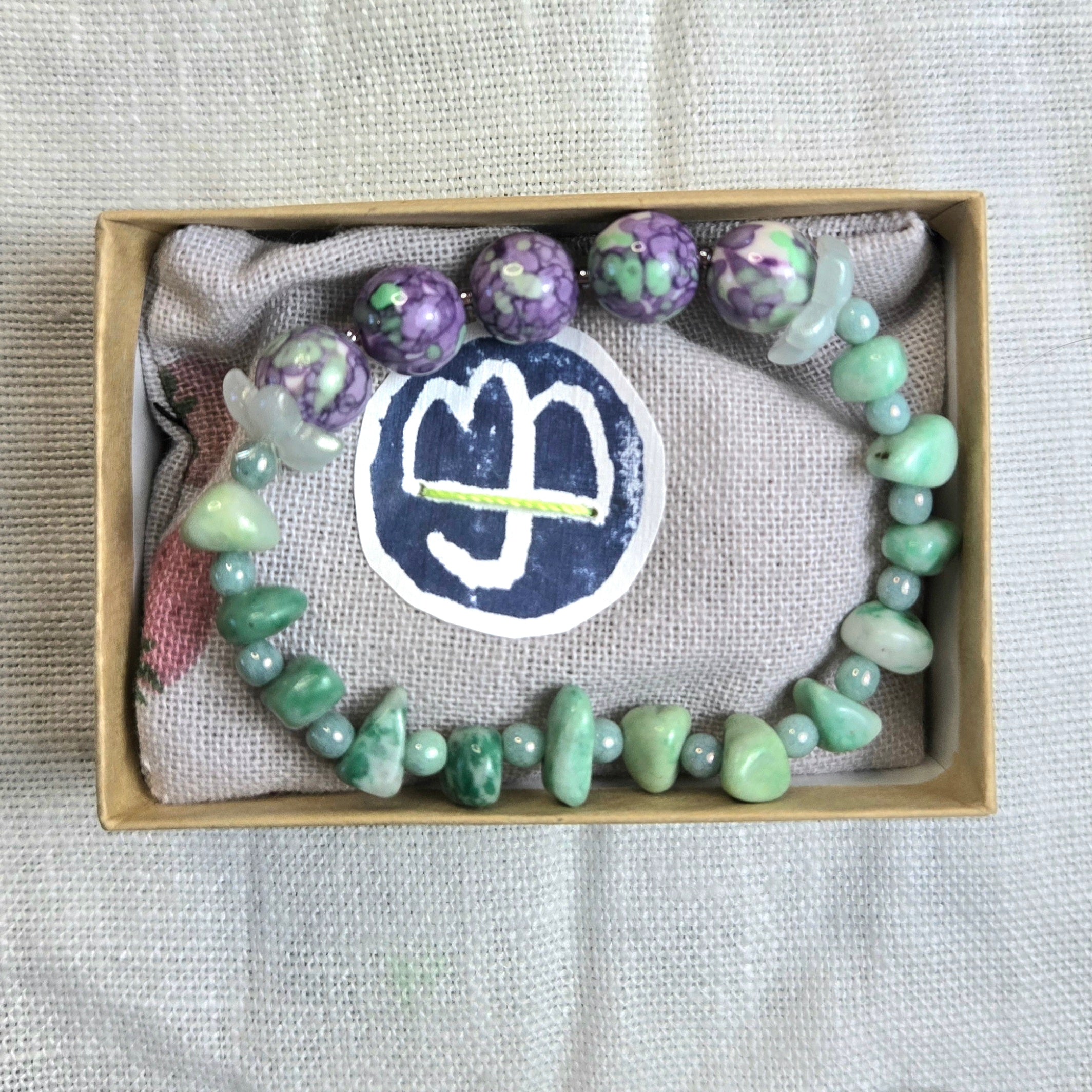 A beaded bracelet displayed inside a kraft gift box lined with textured fabric. The bracelet features light green polished stones, marbled purple and white beads, and a translucent white crystal charm.