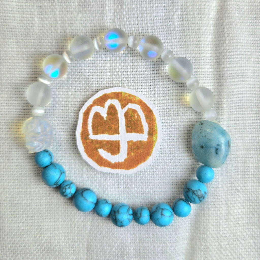 A beaded bracelet arranged in a circle on a textured white fabric background, featuring light blue turquoise beads, milky white beads with iridescent blue flashes, and a large light blue stone. A round white patch with a gold-toned symbol is placed in front of the bracelet.