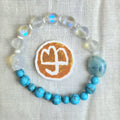 A beaded bracelet arranged in a circle on a textured white fabric background, featuring light blue turquoise beads, milky white beads with iridescent blue flashes, and a large light blue stone. A round white patch with a gold-toned symbol is placed in front of the bracelet.