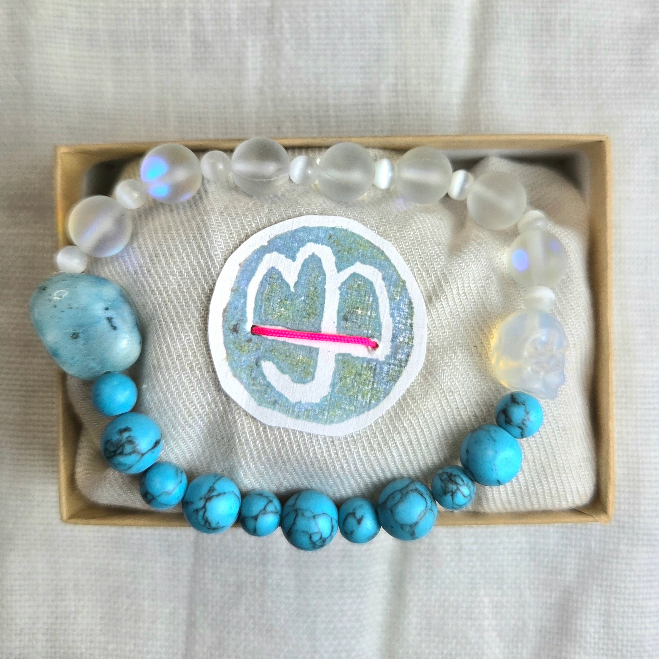 A beaded bracelet displayed inside a light wood gift box on a cream fabric cushion. The bracelet features light blue marbled beads, translucent milky white beads, and a larger textured blue bead.