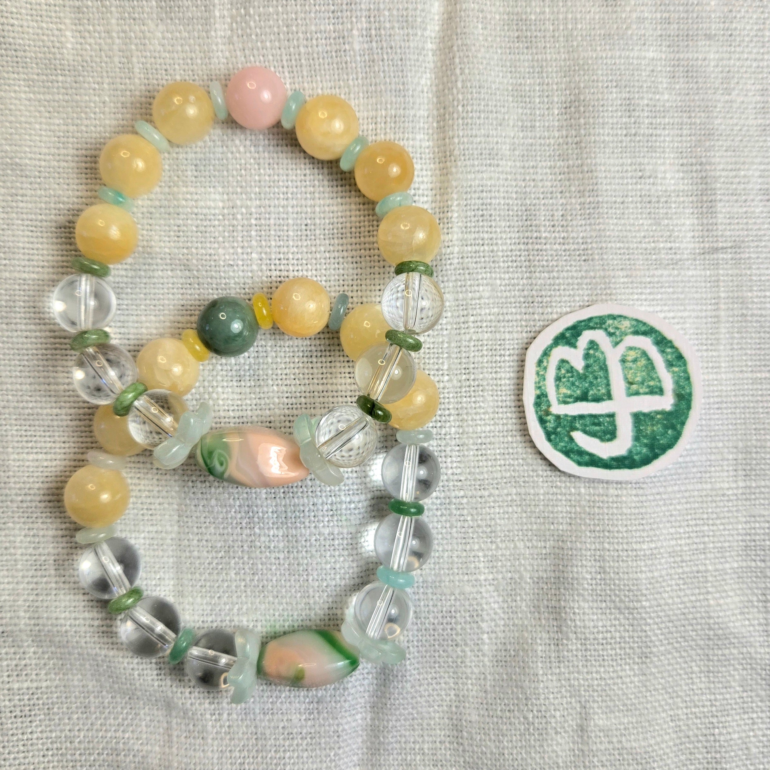 A beaded bracelet laid on a light fabric background, featuring pale yellow beads, clear faceted beads, mint green spacer beads, a pink bead, and a green marbled bead. A small green-and-white round sticker or patch is placed to the right.