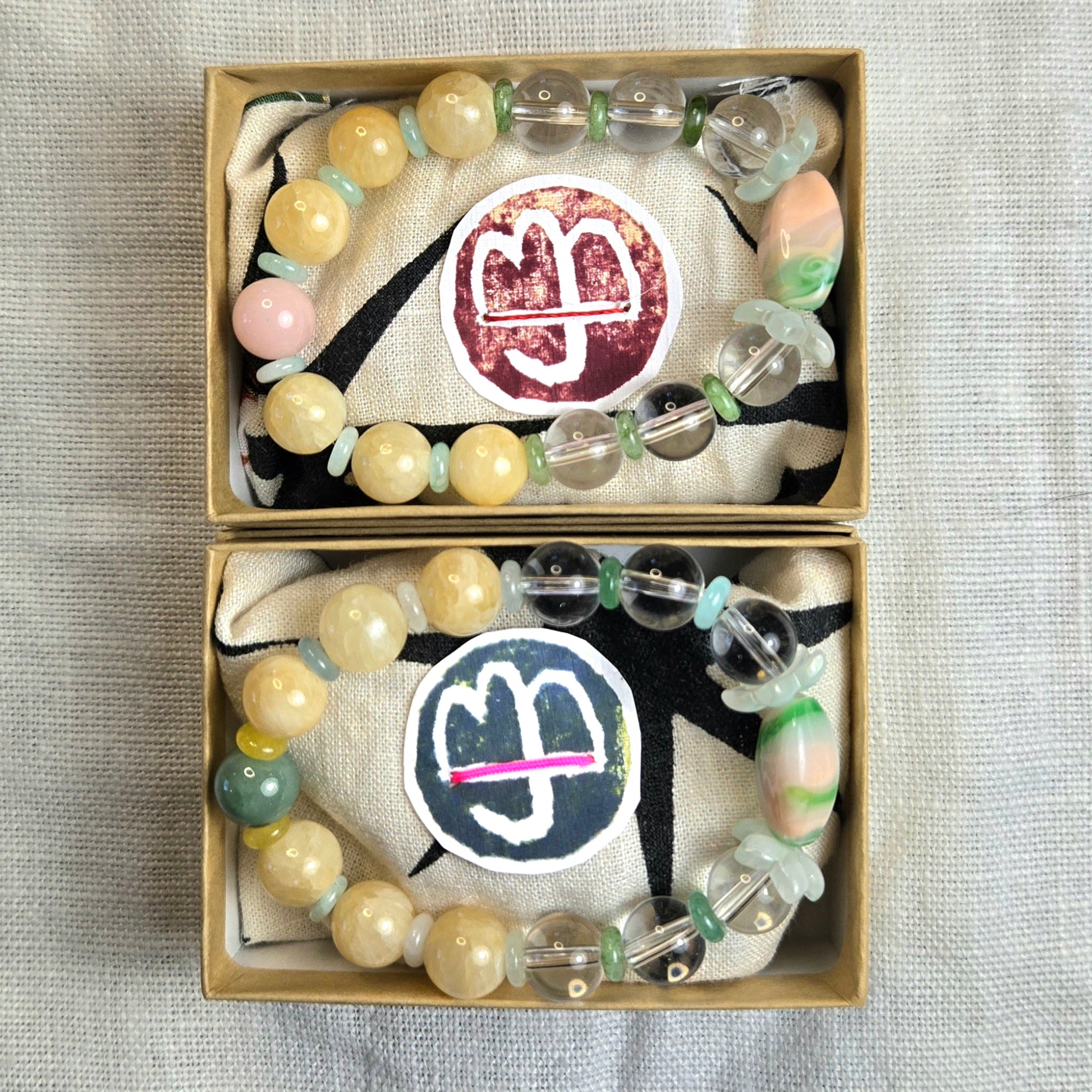 Two beaded bracelets displayed in small kraft gift boxes on a textured fabric surface. The bracelets feature round cream beads, translucent clear beads, pale pink stones, mint green spacer beads, and larger iridescent shell-like accents.