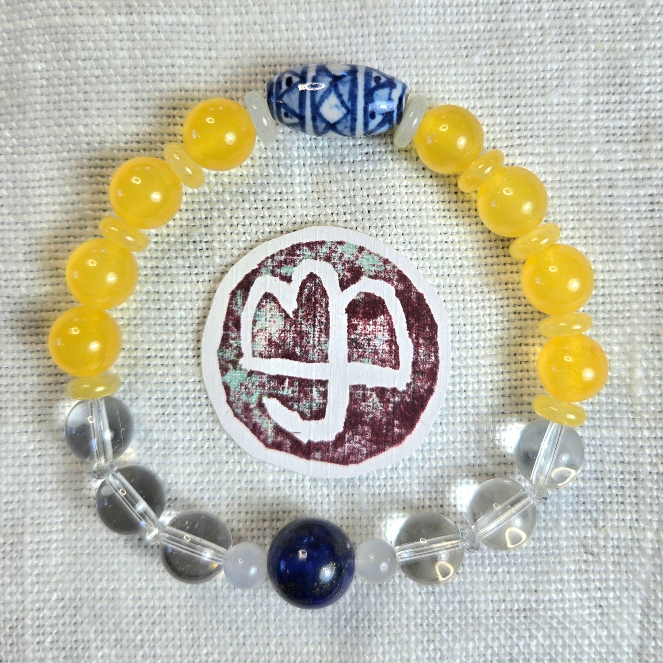 A beaded bracelet arranged in a circle on a textured white fabric background. The bracelet features translucent yellow beads, clear faceted beads, a dark blue round bead, and a blue-and-white patterned accent bead.