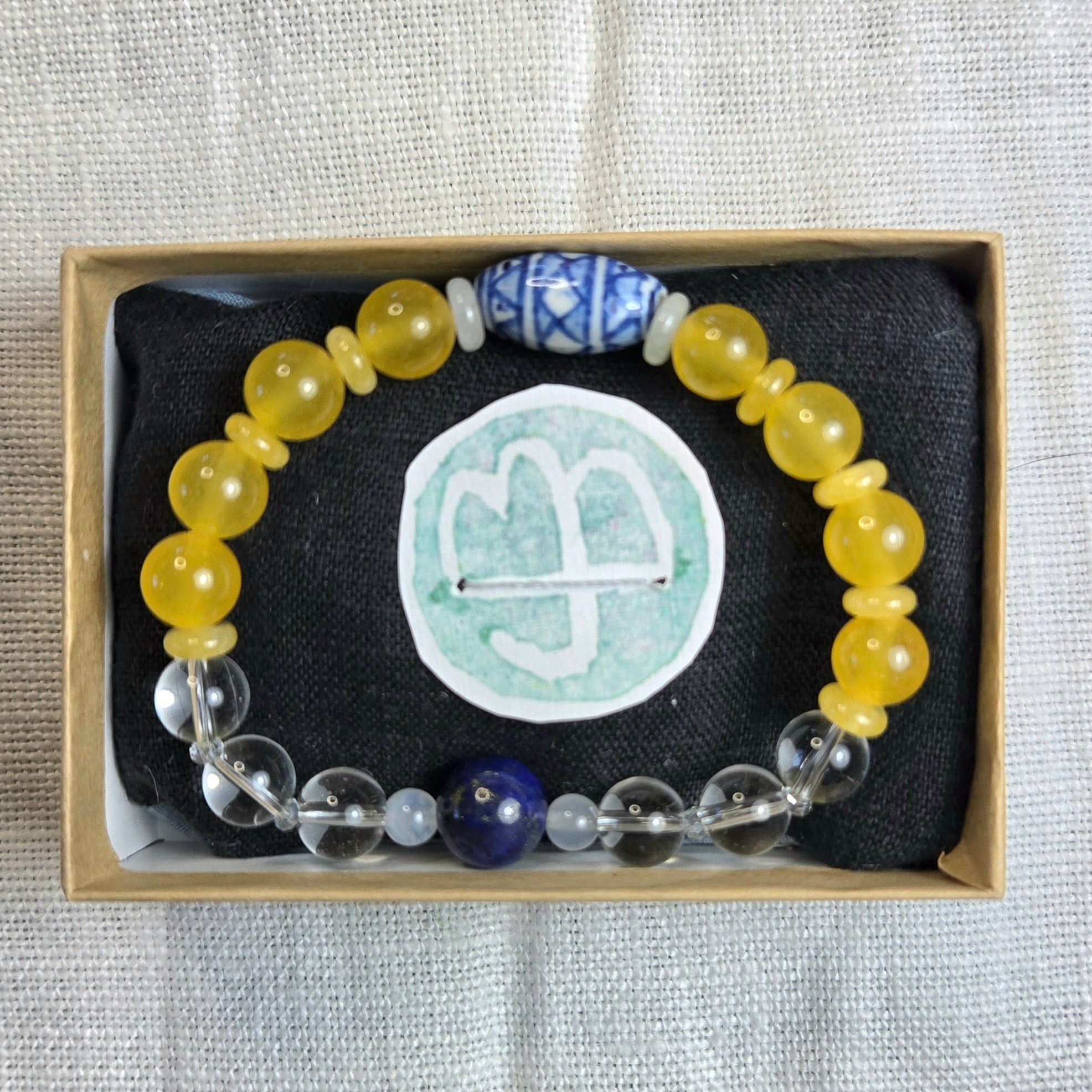A beaded bracelet displayed inside a light wood gift box on black fabric. The bracelet features translucent yellow beads, a blue and white patterned bead, clear beads, and a dark blue bead.