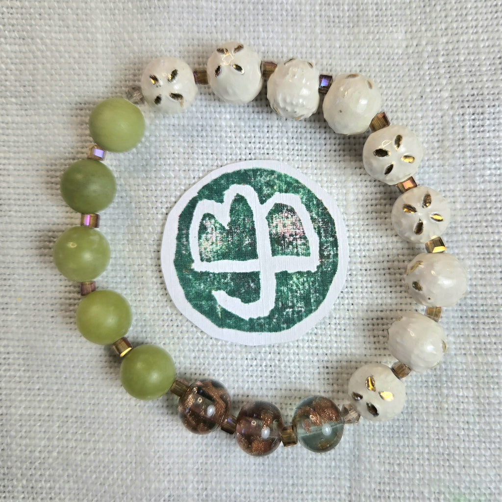 A beaded bracelet arranged in a circle on a textured white fabric background, featuring light green matte beads, white skull-shaped beads with gold accents, and brown translucent beads. A round green-and-white sticker is placed in the center.