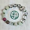 A beaded bracelet arranged in a circle on a white textured fabric background, featuring translucent clear beads, pale green beads, dark green spacer rings, a maroon bead, a brown speckled bead, a white dice-like charm with black markings, and a round green-and-white sticker with a white symbol. No visible brand text or logo is present.