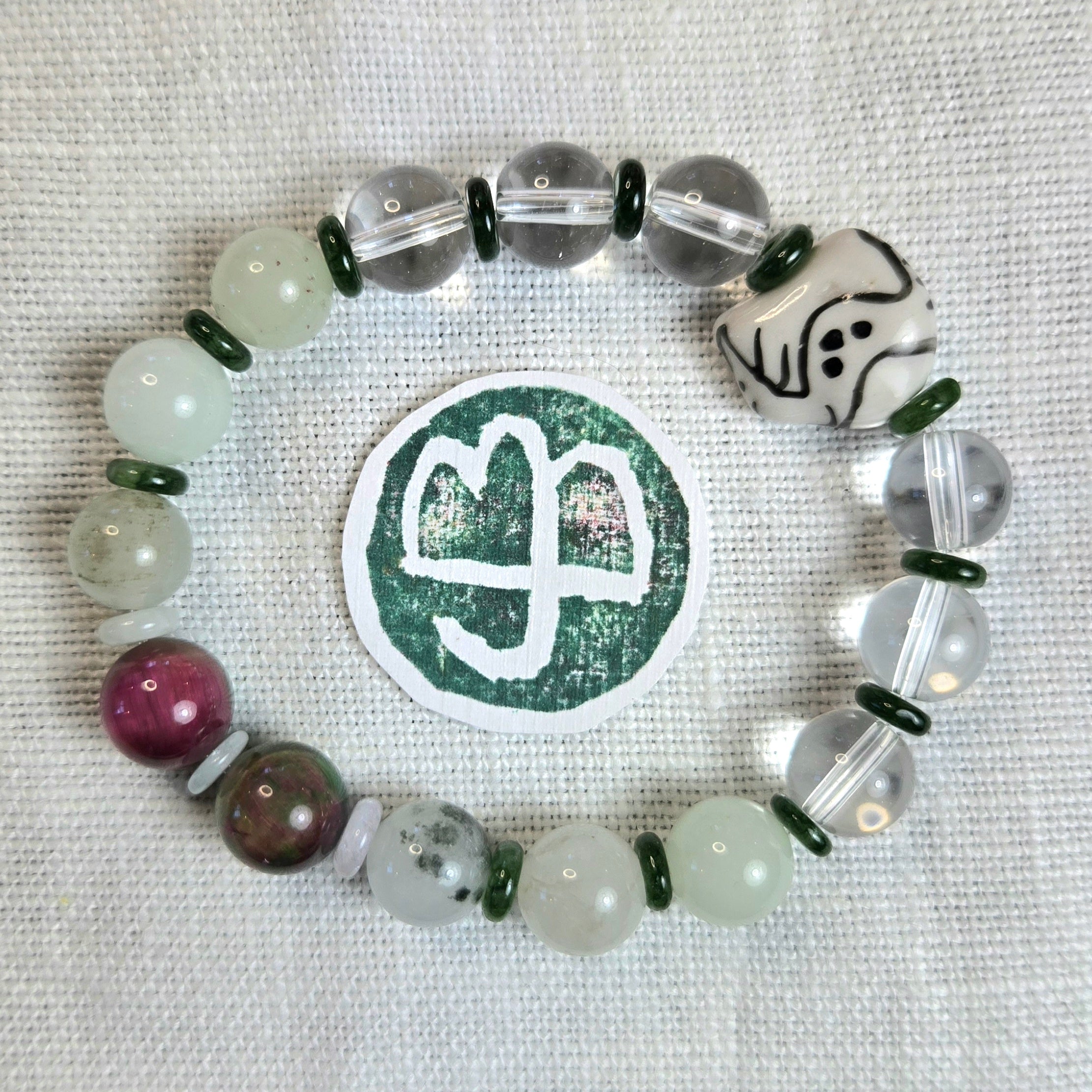 A beaded bracelet arranged in a circle on a white textured fabric background, featuring translucent clear beads, pale green beads, dark green spacer rings, a maroon bead, a brown speckled bead, a white dice-like charm with black markings, and a round green-and-white sticker with a white symbol. No visible brand text or logo is present.