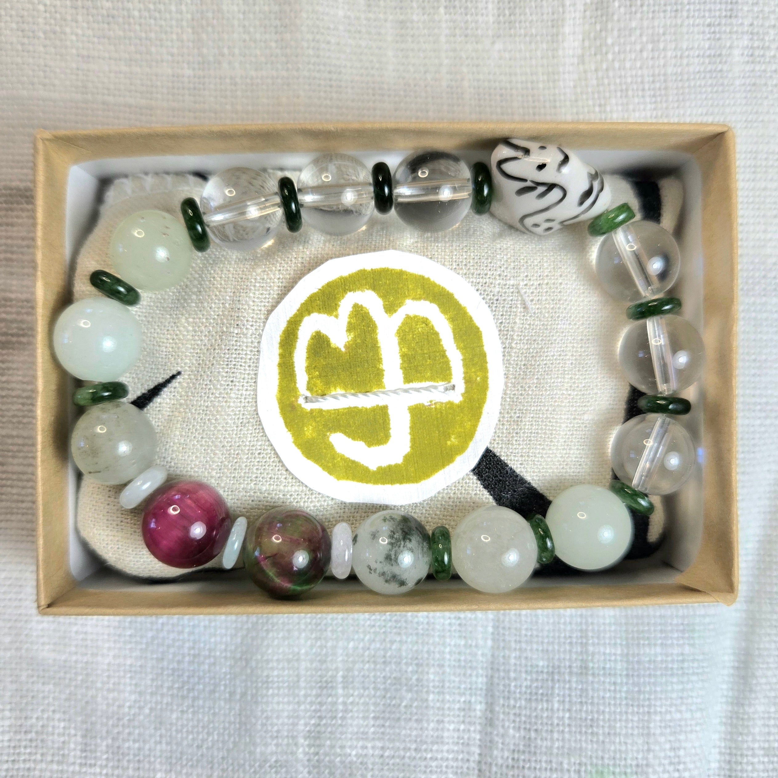 A beaded bracelet displayed inside a light wood gift box on a textured white fabric background. The bracelet features translucent white beads, dark green spacer beads, and polished marbled beads in pink, gray, and white.