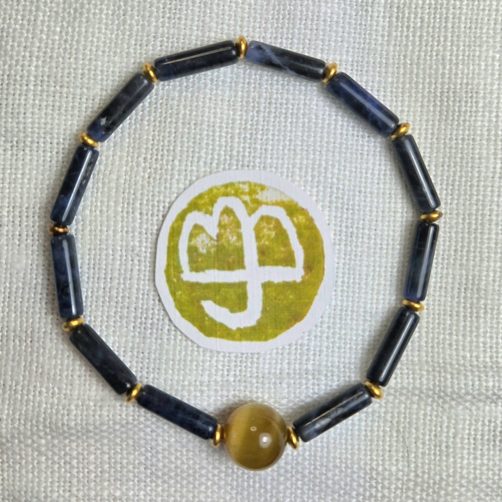 A beaded bracelet made of dark navy blue cylindrical beads separated by small gold spacer rings, with a single round translucent amber-yellow bead at the bottom. The bracelet is photographed on a light beige textured fabric background, with a circular white and olive-green sticker placed in the center.