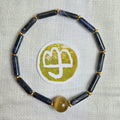 A beaded bracelet made of dark navy blue cylindrical beads separated by small gold spacer rings, with a single round translucent amber-yellow bead at the bottom. The bracelet is photographed on a light beige textured fabric background, with a circular white and olive-green sticker placed in the center.