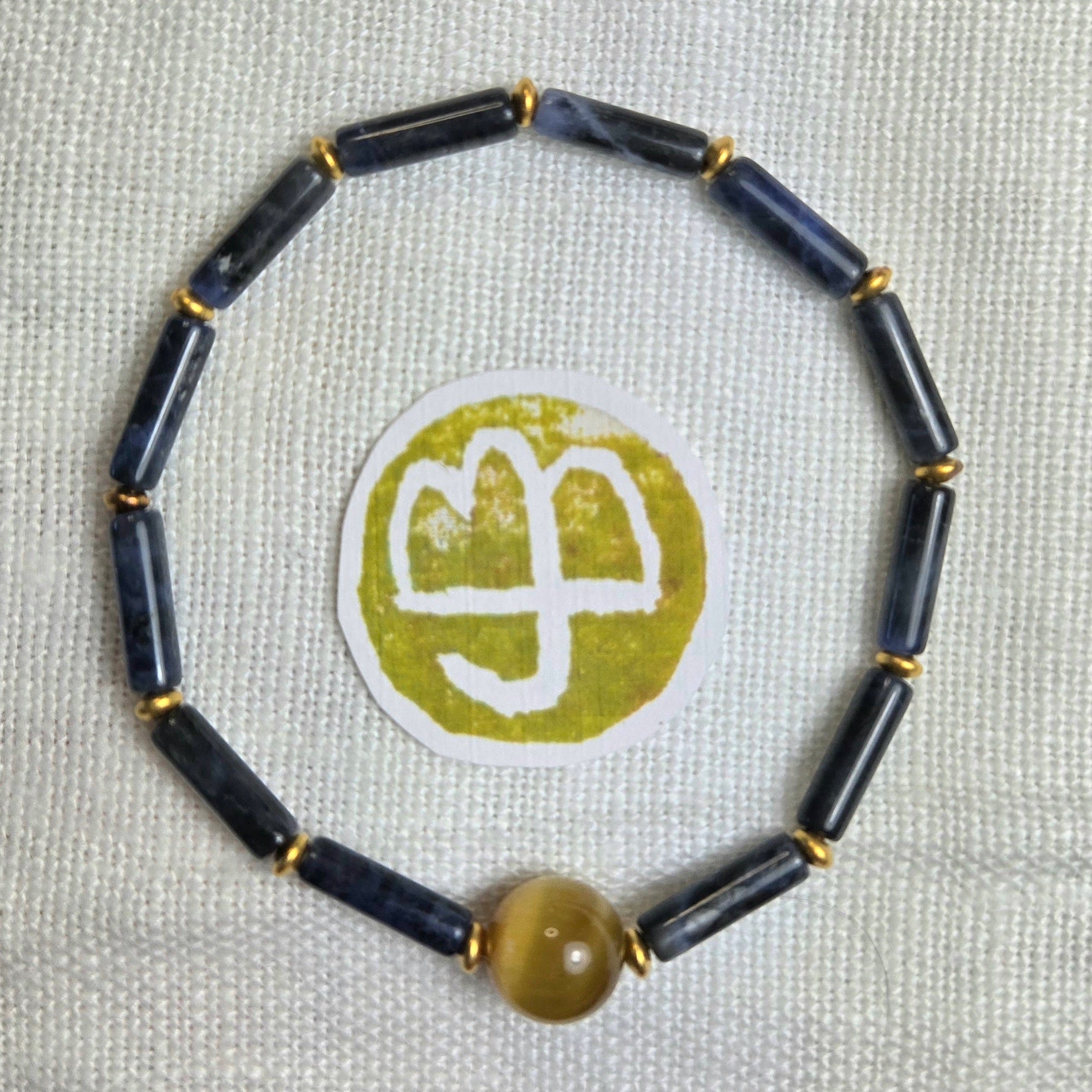 A beaded bracelet made of dark navy blue cylindrical beads separated by small gold spacer rings, with a single round translucent amber-yellow bead at the bottom. The bracelet is photographed on a light beige textured fabric background, with a circular white and olive-green sticker placed in the center.